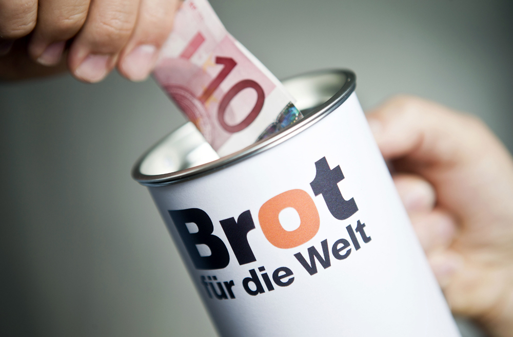 Spendendose mit Aufschrift „Brot für die Welt“, jemand wirft 10-Euro-Schein hinein