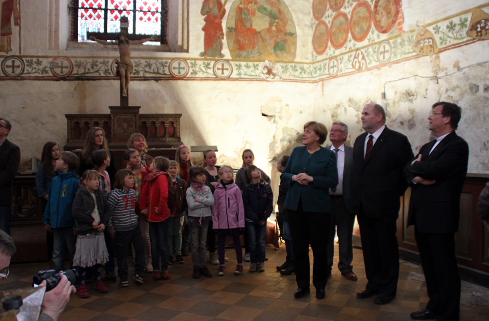 Kanzlerin Merkel begutachtet die Wandmalereien, während die Kinder ein Ständchen singen