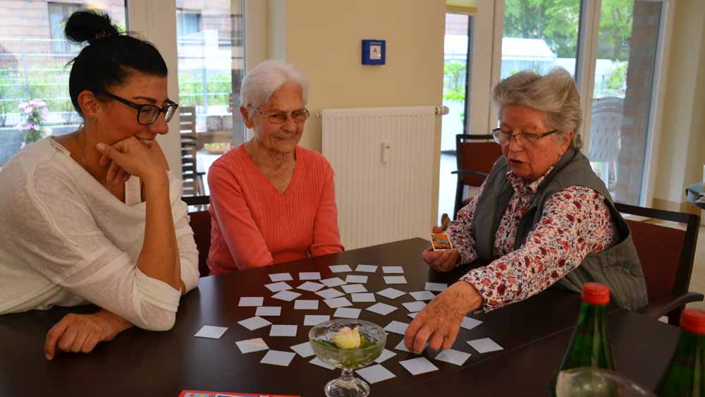 Pflegerin Conny Rowe und die Patientinnen Anneliese und Ingrid sitzen an einem großen Tisch und spielen Memory.