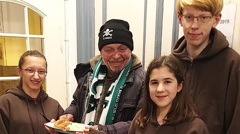 Drei junge Helfer reichen einem Gast eine Mahlzeit