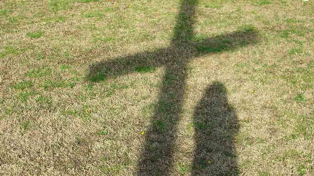 Schatten eines Kreuzes auf einem Rasen, daneben Schatten eines Menschen