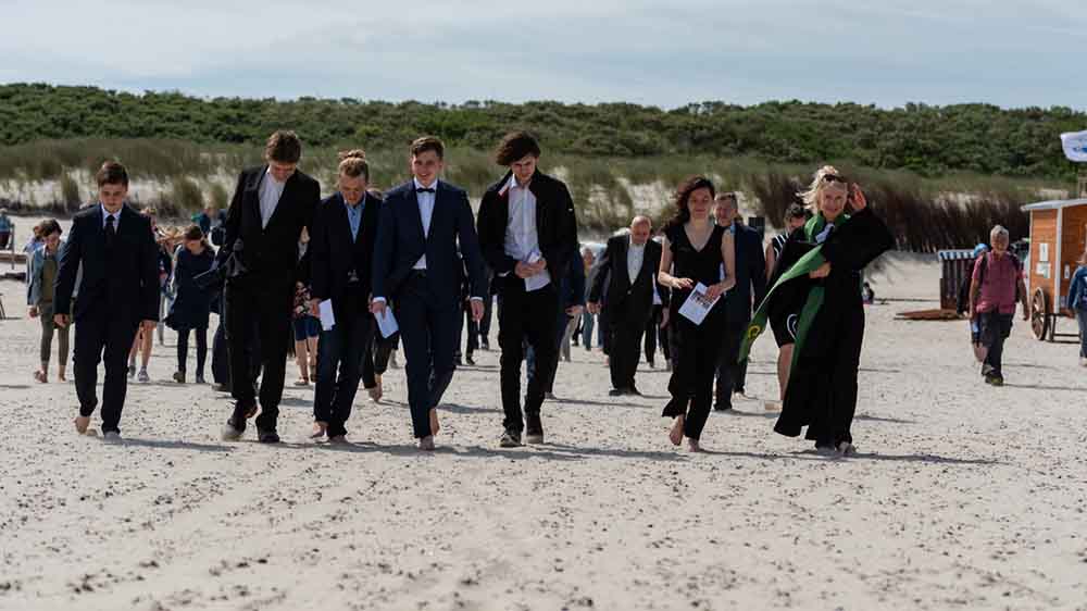 Konfis im Anzug und Pastorin marschieren durch den Sand