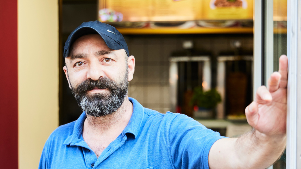 Cervat Mergen vor seinem Restaurant “Toros” in Osnabrueck (Foto vom 04.08.2021). Neuerdings serviert er zu Kebap, Pommes und Salat auch Gedichtverse. Das Berliner Kuenstlerkollektiv Kasia Korczak und Payam Sharifi, genannt “Slavs und Tatars”, hat Servietten, Doenertueten, Bierdeckel und mobile Holzaufsteller in dem Doenerladen mit Versen aus einem tuerkischen Epos aus dem 11. Jahrhundert bedrucken lassen. Stammgaeste und kunstinteressierte neue Gaeste koennen sie waehrend des Essens lesen und darueber miteinander ins Gespraech kommen. Mergen und seine Stammgaeste freut es. Fast jeden Tag kommen neue, kunstinteressierte Gaeste dazu. An manchen Tagen gibt es ausserdem Lesungen, Performances oder Podcasts zu hoeren und zu sehen. Das Projekt “Asbildung” (As ? tuerkisch fuer “warme Speise”) entstand in Zusammenarbeit mit der Kunsthalle Osnabrueck. (Siehe epd-Bericht vom 16.08.2021)