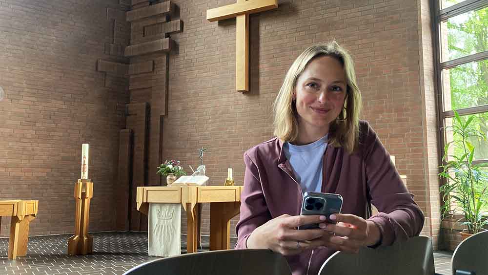 Teske sitzt auf Kirchenbank und lehnt sich mit Smartphone in der Hand zur Kamera, im Hintergrund der Altar