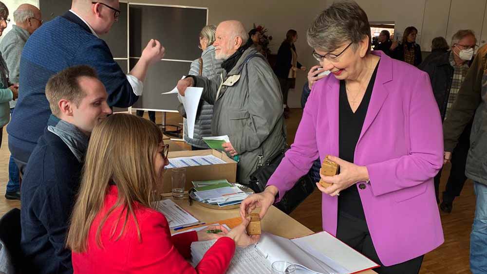 Kühnbaum-Schmidt im Wahllokal, verteilt kleine Pakete an Helfer, die am Tisch sitzen