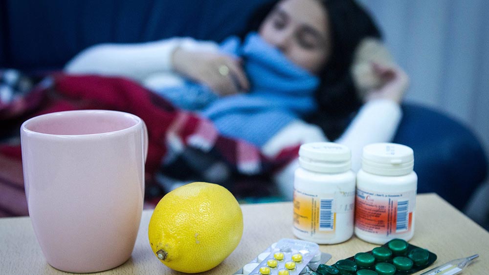 Frau liegt mit dickem Schal im Bett, davor Medikamente und eine Tasse