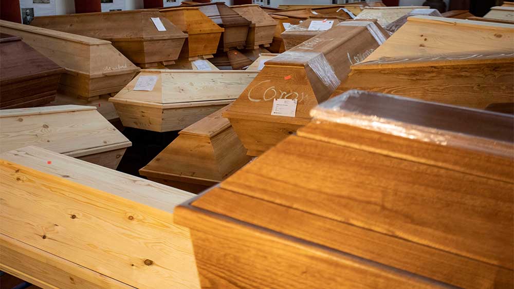 Braune Holzsärge stehen in einem Krematorium. An vielen hängt ein Zettel mit der Aufschrift "Infektionsgefahr".