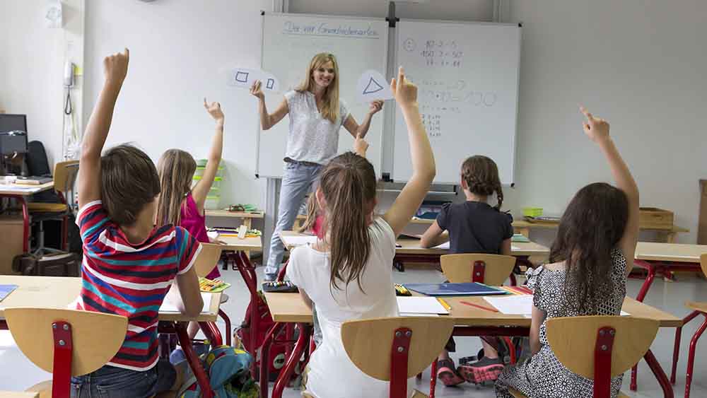 Lehrerin steht vor Klasse im Klassenraum, viele Kinder melden sich
