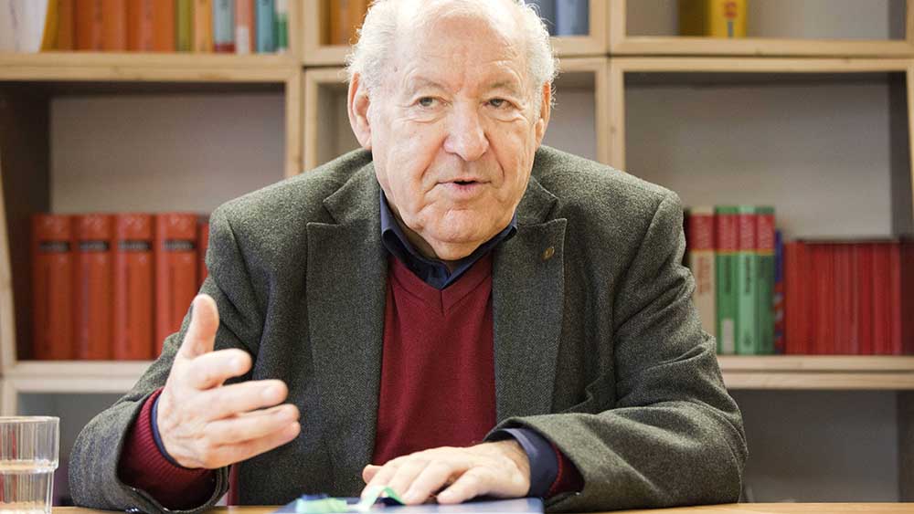 Perel im braunen Sakko vor einem Bücherregal mit Buch in der Hand