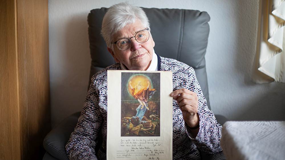 Eine ältere Dame sitzt im Sessel und hält ihre Konfirmationsurkunde in die Kamera.