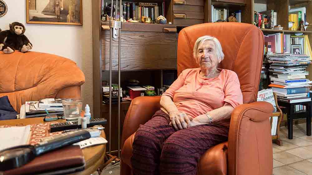 Alte Frau sitzt im Wohnzimmer im Sessel, große Regalwand im Hintergrund