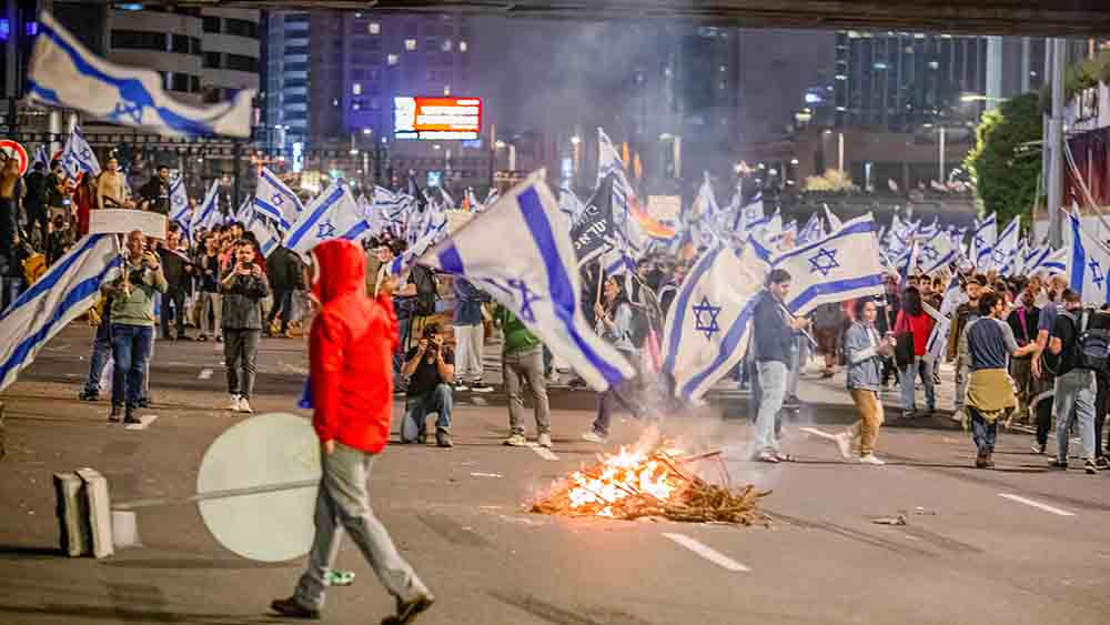 Menschen tragen viel Israel-Flaggen durch eine Straße, ein kleines Feuer brennt