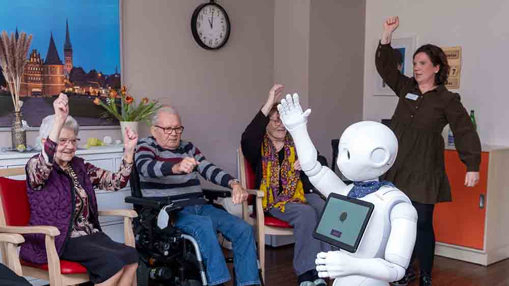 Weißer Roboter hebt die Arme, zwei Senioren sitzen dahinter und schauen zu