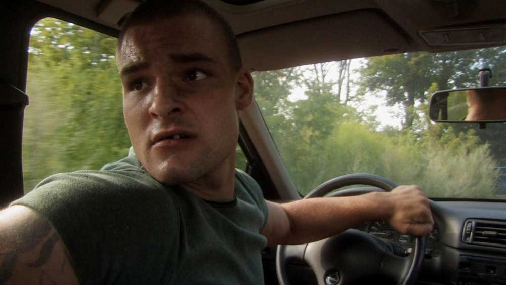 Ein Ausschnitt aus dem Filmplakat. Ein junger Mann sitzt mit nervösem Blick im Auto