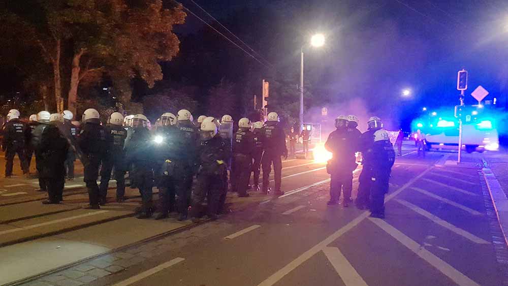 Polizisten in Kampfmontur laufen eine Straße entlang, im Hintergrund brennt ein Feuer