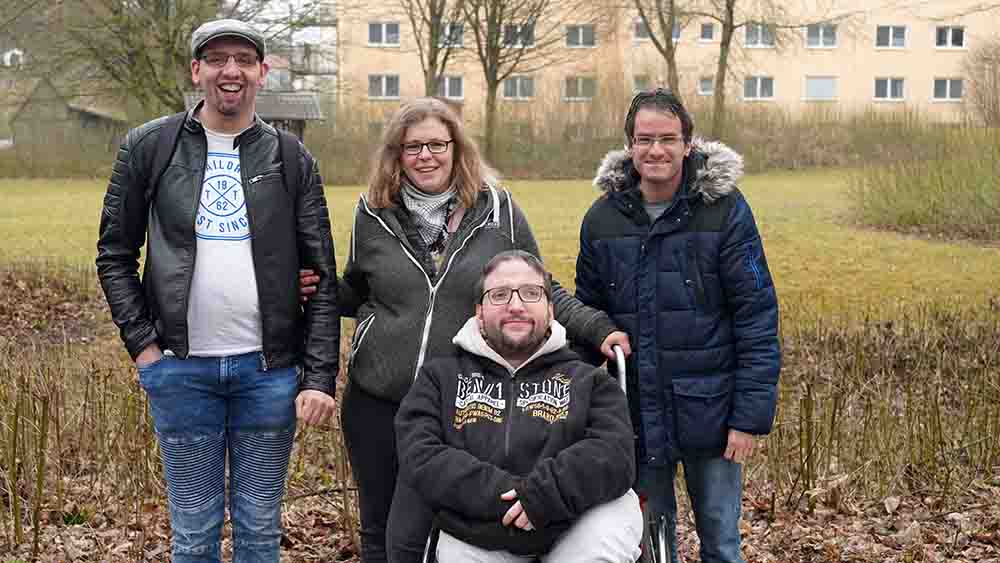 Drei Männer, eine Frau auf einer Wiese. Einer der Männer sitzt in einem Rollstuhl