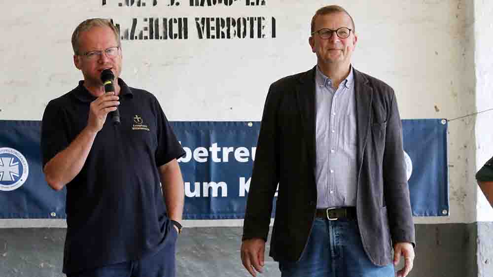Zwei Männer stehen auf einer kleinen Bühne, der linke Mann spricht in ein Mikro