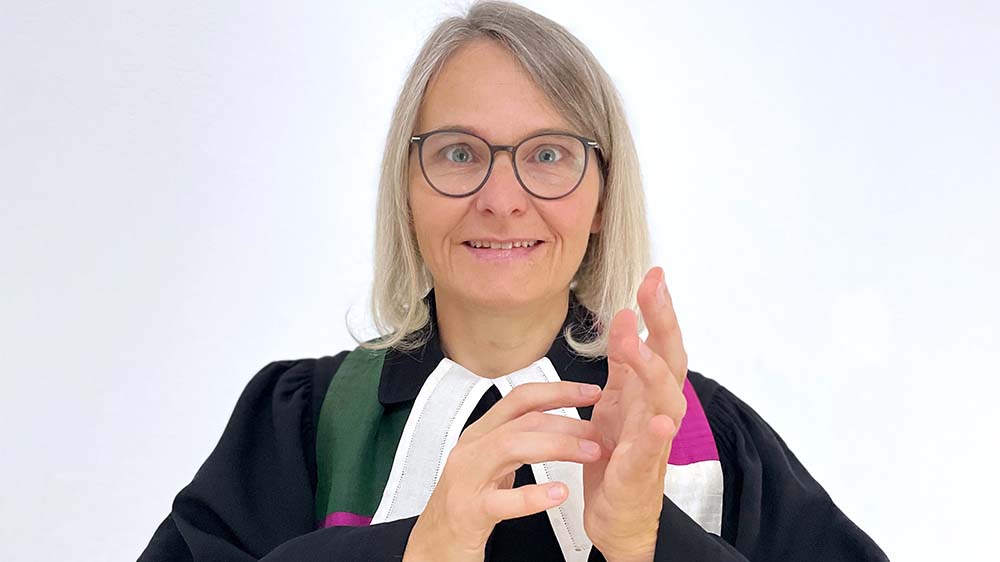 Mittelalte Frau mit blonden Haaren und Brille in Talar gebärdet das Wort "Jesus"