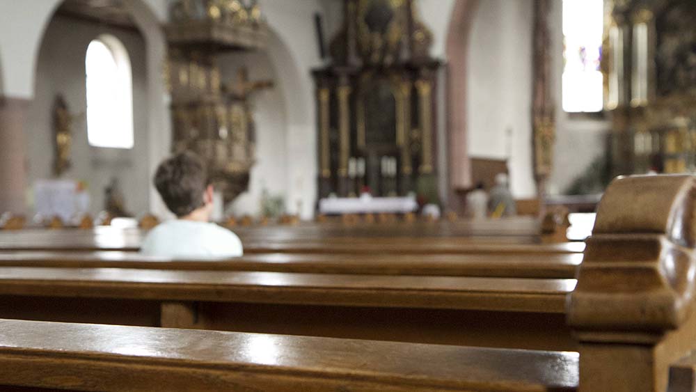 Eine Person, von hinten fotografiert, sitzt auf einer Kirchenbank. Die anderen Plätze sind leer