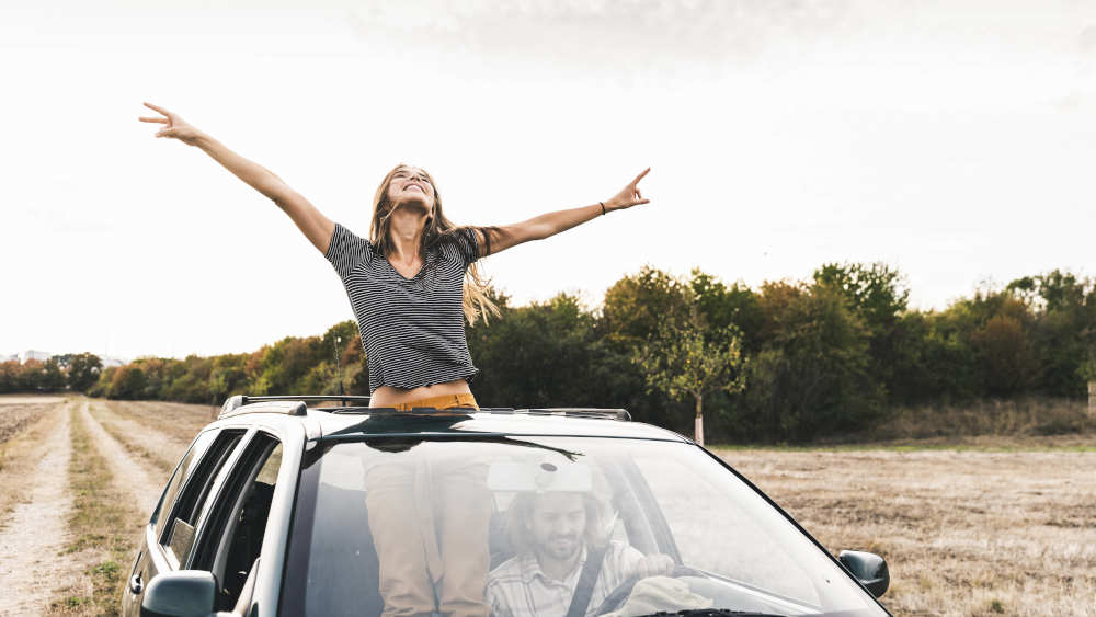 Junge Frau streckt glücklich ihre Arme aus einem fahrenden Auto