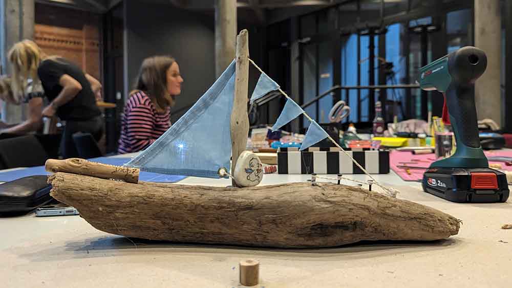 Kleines Schiff aus Holz mit Segel steht auf einem Tisch, daneben eine Bohrmaschine
