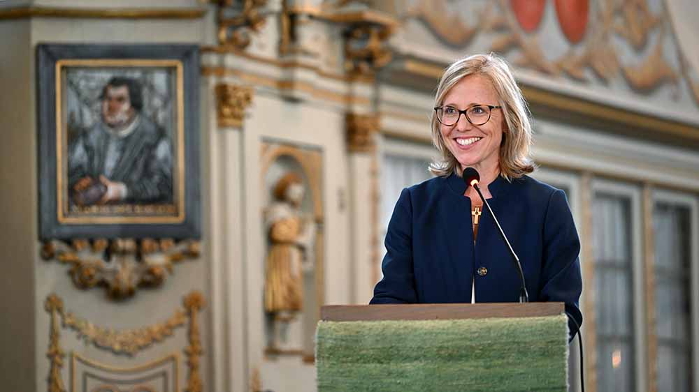 Blonde Frau mit Brille und dunkelblauem Blazer steht am Rednerpult einer Kirche