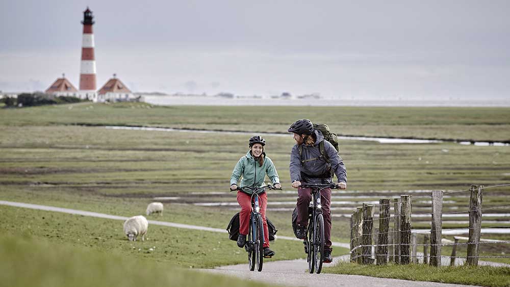 Zwei Menschen fahren mit Fahrrädern über grüne Wiesen, im Hintergrund ein Leuchtturm