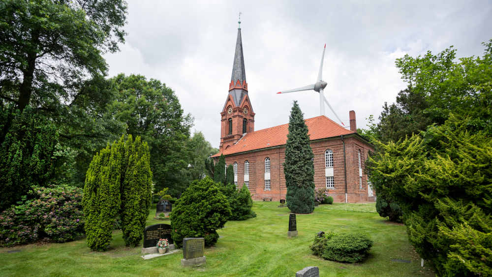 Kirche und Friedhof stehen auf einer Grünfläche mit altem Baumbestand an der Autobahn A7, einige hundert Meter von der Autobahnauffahrt entfernt