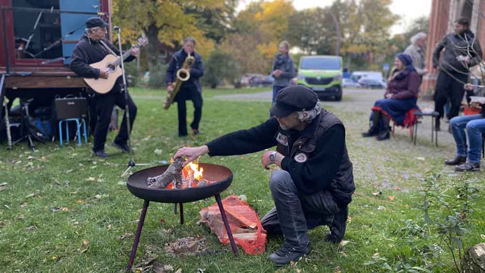 Mann hockt sich hin und macht Feuer in einer Feuerschale, im Hintergrund spielen zwei Leute Saxofon und Flöte