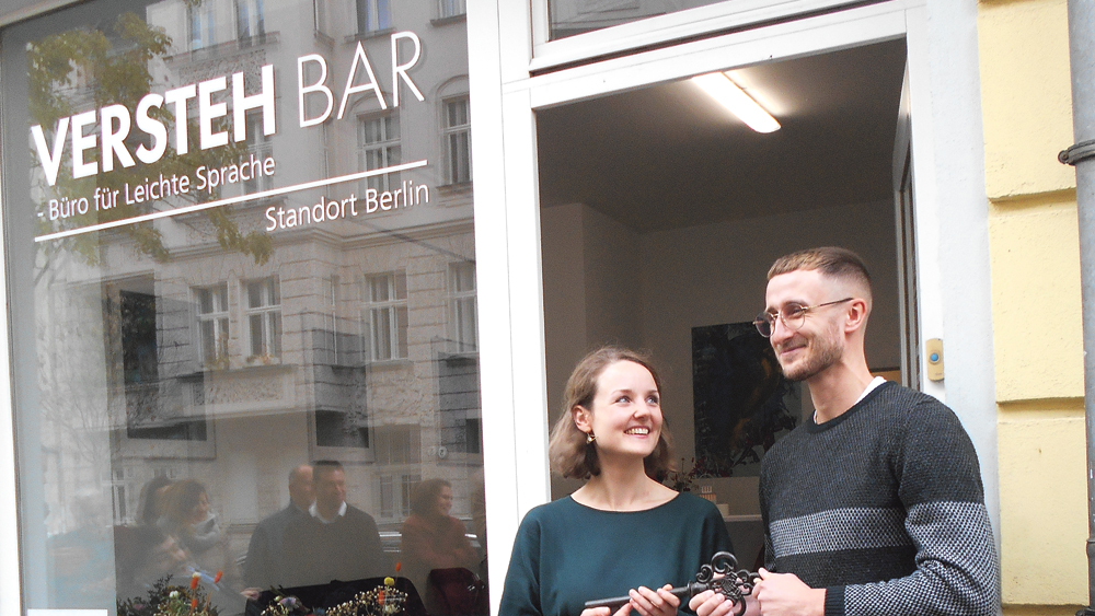 Ein Mann und eine Frau vor einem Gebäude. Auf der Fensterscheibe steht "Versteh-Bar"