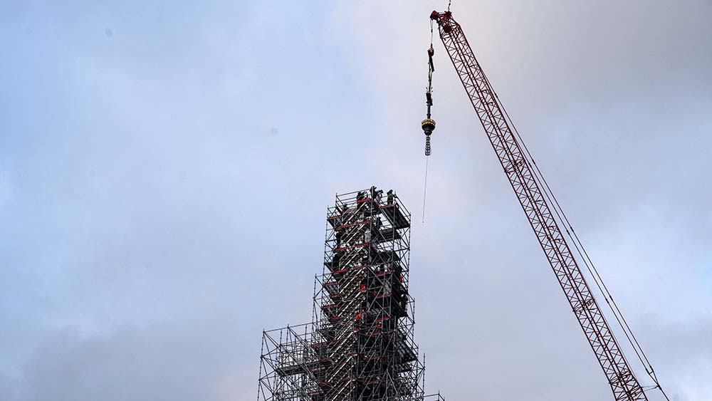 An einem roten Kran hängt ein goldenes Kreuz, das auf einen eingerüsteten Turm gesetzt wird