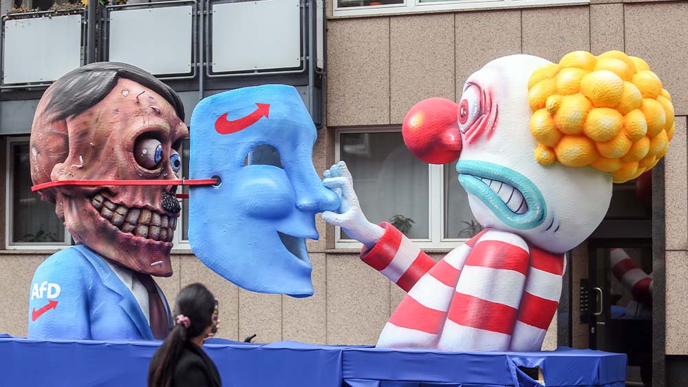 Ein kostümierter Narr reißt einer AfD-Figur eine Maske vom Gesicht, hinter der eine tiefbraune Fratze sichtbar wird, die an Hitler erinnert