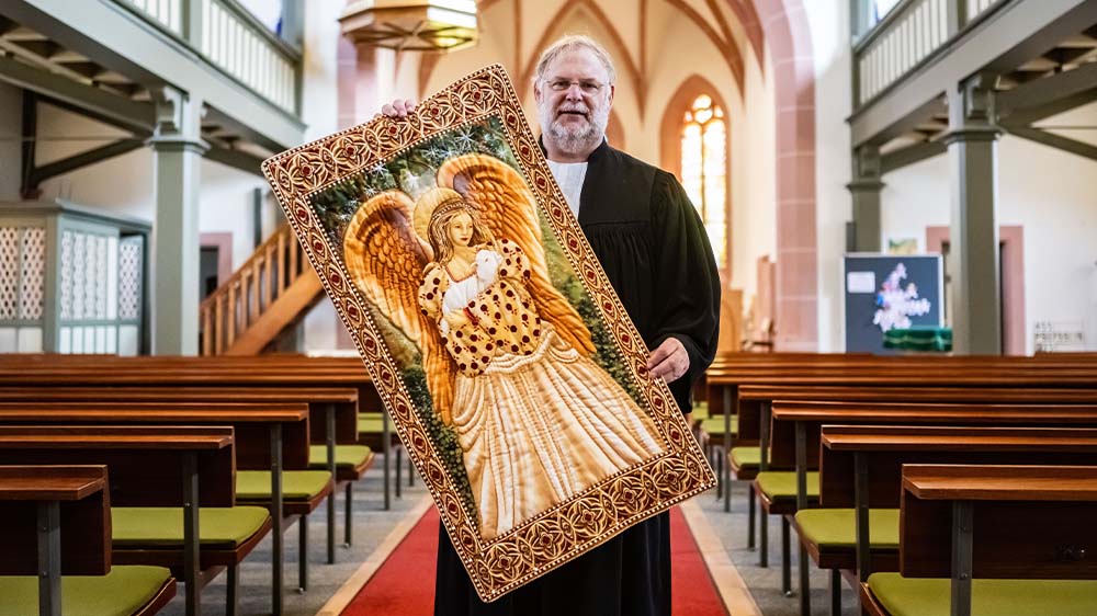 Pfarrer Jörg Seiter in der evangelischen Kirche der Michaelisgemeinde Blankenloch mit einem fertigen Wandbehang "Engelsquilt"