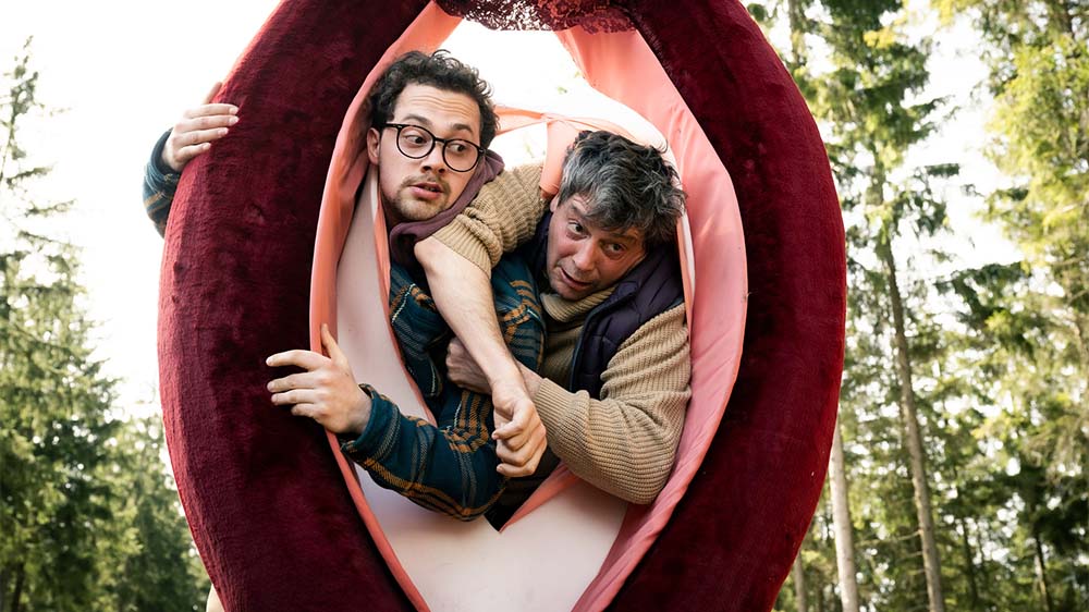 "Überväter": Luca (Anselm Bresgott) und Mathi (Fritz Karl) versuchen beide gleichzeitig durch eine überlebensgroße Vulva zu klettern und blockieren sich gegenseitig