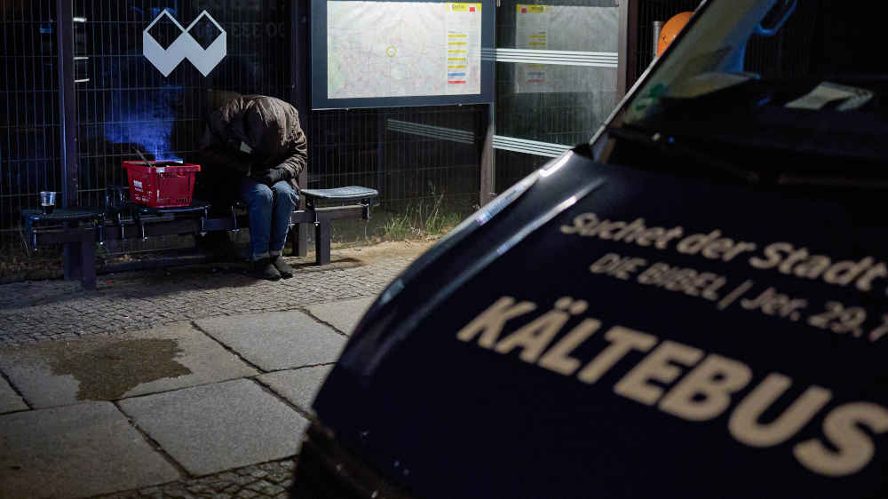 Ein Kältebus steht vor einer Bushaltestelle, an der eine obdachlose Frau sitzt