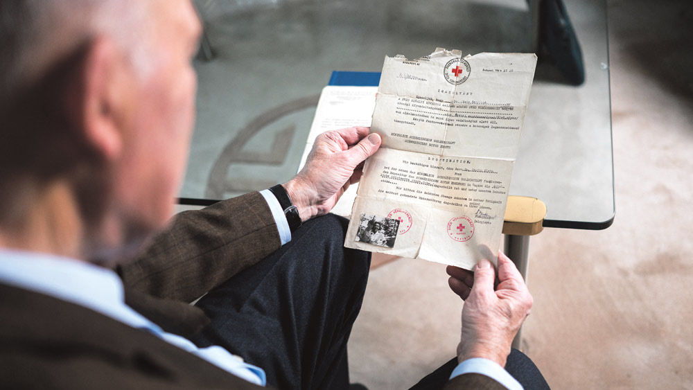 Der Holocaust-Überlebende Peter Kenedi sitzt auf einer Couch in seinem Wohnzimmer in Frankfurt am Main. In den Händen hält er einen ungarischen Schutzpass des Roten Kreuzes, darauf ein Foto, das ihn im Alter von sechs Jahren zusammen mit seiner Mutter Susanna zeigt. Peter Kenedi überlebte als Kind das Ghetto Budapest