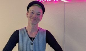 Ein Mensch steht vor einem pinken Leuchtschild mit der Aufschrift "God ist queer"