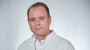 Ein Mann im weißen Kittel schaut in die Kamera. Der Hintergrund ist ebenfalls weiß.