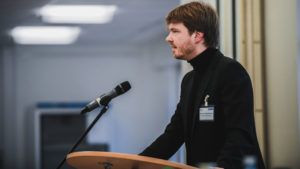 Mann mit dunklen Haaren, Vollbart und schwarzem Pullover an einem Rednerpult