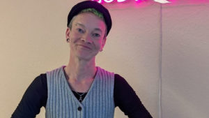 Ein Mensch steht vor einem pinken Leuchtschild mit der Aufschrift "God ist queer"